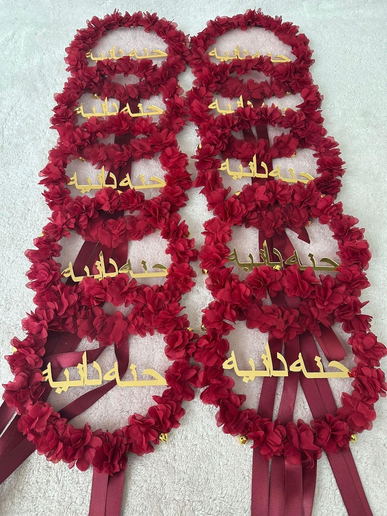 May include: A collection of burgundy floral wreaths with gold lettering. Each wreath is adorned with small, fabric flowers and long, burgundy ribbons. The gold lettering appears to be Arabic script. The wreaths are likely for a special occasion.