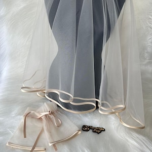May include: A sheer white bridal veil with a delicate gold trim, draped over a mannequin. Two matching wrist cuffs with gold trim and ribbon ties are also visible. The items are displayed on a fluffy white surface.