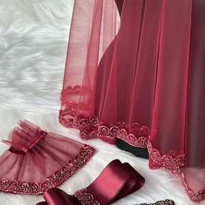 Pu&ograve; includere: Un velo di tulle color borgogna con un bordo di perline, una fascia di raso color borgogna abbinata con un bordo di perline e un braccialetto di tulle color borgogna con un bordo di perline. Gli articoli sono su una superficie bianca e soffice.