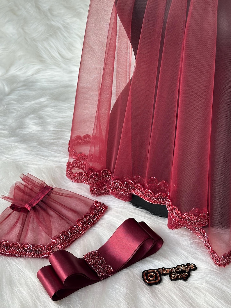 Pu&ograve; includere: Un velo di tulle color borgogna con un bordo di perline, una fascia di raso color borgogna abbinata con un bordo di perline e un braccialetto di tulle color borgogna con un bordo di perline. Gli articoli sono su una superficie bianca e soffice.