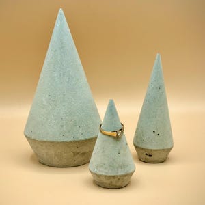 Könnte beinhalten: Drei kegelförmige Ringhalter in verschiedenen Größen. Die Halter sind hellblau mit einer gesprenkelten Textur und einem Sockel in Betonfarbe. Ein goldener Ring mit einem Diamanten wird auf dem kleinsten Halter präsentiert. Der Hintergrund ist einfarbig beige.