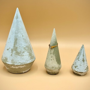 May include: Three concrete ring holders in varying sizes. The largest is a tall cone shape, the middle one holds a gold ring, and the smallest is a miniature cone. All are painted with a distressed white and tan finish.