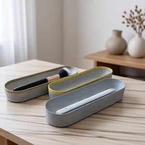 Puede incluir: Tres bandejas ovaladas de hormigón con interiores grises lisos y bordes contrastantes en amarillo o gris. Una bandeja contiene una brocha de maquillaje negra y dorada, mientras que otra contiene un lápiz óptico blanco. Diseño bajo.