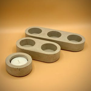 Puede incluir: Tres candelabros de hormigón de diferentes tamaños. Dos son oblongos con huecos circulares para velas, uno con tres y otro con dos. También hay un soporte redondo más pequeño con una vela blanca encendida.