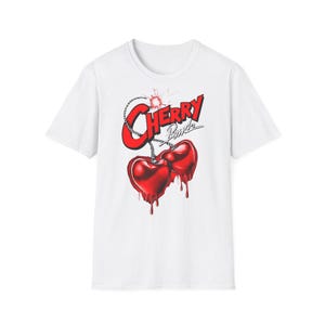 Puede incluir: Camiseta blanca con un diseño gráfico. El diseño presenta el texto "Cherry Bomb" en rojo, encima de dos cerezas rojas en forma de corazón con detalles goteantes. Una cuerda y chispas rojas completan el diseño.