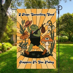 May include: A yellow and orange garden flag with a cartoon frog wearing a hat and carrying a bag. The frog is walking on a path with mushrooms and flowers. The text on the flag reads "I hope something good happens to you today."