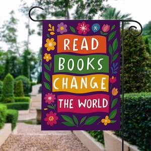 May include: A purple garden flag with the words "READ BOOKS CHANGE THE WORLD" in white, set against orange, green, yellow, and red rectangular backgrounds. The flag is decorated with colorful flowers and green leaves.