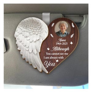 Peut inclure: Un charm de voiture commémoratif en forme de cœur avec une photo d'une femme, le nom "Anna" et les dates 1964-2025. Le charme a un aspect boisé avec le texte "Although You cannot see me I am always with You". Une aile d'ange blanche est attachée.