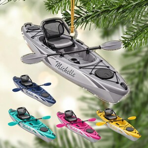 Könnte beinhalten: Ein graues Kajak-Ornament mit schwarzem Sitz, Doppelpaddel und schwarzer Bungee-Seil-Befestigung. Die Seite des Kajaks ist mit dem Namen Michelle in Schreibschrift personalisiert. Weitere Kajak-Ornamente sind in Blau, Türkis, Pink und Gelb abgebildet.