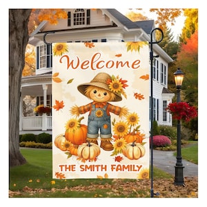 Puede incluir: Una bandera de jardín con un diseño de espantapájaros con girasoles, calabazas y hojas de otoño. La bandera dice "Welcome" y "The Smith Family". El espantapájaros lleva un sombrero de paja y un peto, creando una bienvenida con temática otoñal.