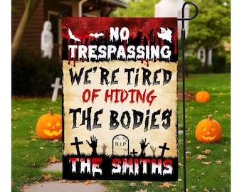 May include: Halloween garden flag with the text "NO TRESPASSING WE'RE TIRED OF HIDING THE BODIES THE SMITHS" in red and white. The flag has a spooky design with bats, a tombstone, and zombie hands. Two carved pumpkins and a ghost decoration are in the background.