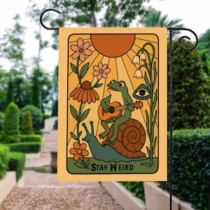 Puede incluir: Una bandera de jardín amarilla con una rana verde tocando la guitarra sobre un caracol. La bandera tiene un sol, flores y el texto "Stay Weird".