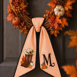 May include: A seasonal wreath featuring orange maple leaves, berries, pinecones, and pumpkins, accented by a hanging orange and white striped fabric ribbon. The ribbon displays a graphic of brown cowboy boots with a sunflower and the letter M.