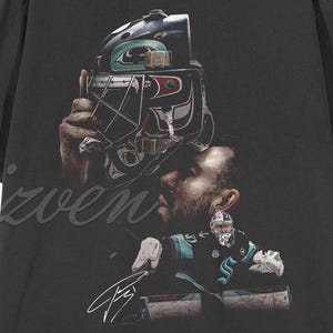 Puede incluir: Camiseta negra con un gráfico detallado de una máscara de portero de hockey y un jugador con uniforme. El diseño incluye una firma y el logotipo del equipo. La camiseta está hecha de un material suave y cómodo.