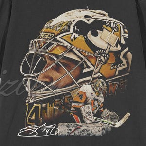 May include: Black t-shirt featuring a detailed graphic of a hockey goalie mask in gold and black, with the number 74 and a signature. The design includes a cityscape and hockey equipment.