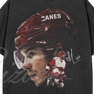 Könnte beinhalten: Ein anthrazitfarbenes Kurzarm-T-Shirt mit einem großen Grafikdruck eines Eishockeyspielers, der einen roten Helm mit dem Text CANES trägt. Das Design zeigt ein Nahaufnahme-Gesicht des Spielers, eine Ganzkörper-Actionaufnahme und eine handschriftliche Signatur mit 37.