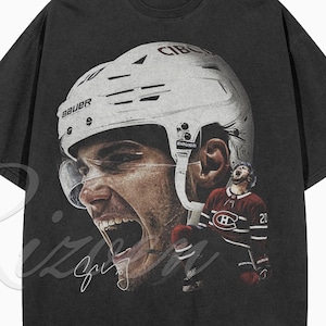 Könnte beinhalten: Ein anthrazitfarbenes Kurzarm-T-Shirt mit einem großen Grafikdruck eines Eishockeyspielers, der einen weißen Bauer-Helm trägt. Das Design zeigt eine Nahaufnahme des Gesichts, eine Ganzkörperansicht des Spielers im roten Trikot und eine Signatur.