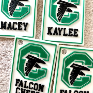 Op de afbeelding: Witte rechthoekige sleutelhangers met een groen en zwart valklogo en tekst. De sleutelhangers tonen de namen "Macey" en "Kaylee", en de tekst "Falcon Cheer". De sleutelhangers hebben een groene rand.