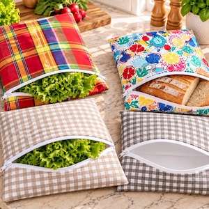 Peut inclure: Quatre sacs de rangement carrés en tissu avec fermetures éclair blanches. Un sac présente un motif à carreaux rouge, jaune et bleu contenant de la laitue. Un autre a un imprimé d'agrumes colorés contenant du pain tranché. Deux sacs ont des motifs vichy, un beige et un gris, dont l'un contient de la laitue.