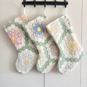 May include: Three quilted Christmas stockings hanging from a black metal rack. Each stocking features a patchwork design with floral patterns in pastel shades of pink, blue, yellow, and green, constructed from hexagonal fabric pieces.