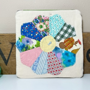 May include: A white fabric pillow with a patchwork flower design. The flower is made of different colored fabrics with floral and geometric patterns. The center of the flower is a yellow circle.