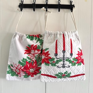 May include: Two white drawstring bags with Christmas-themed designs. One features red poinsettias and green holly, while the other showcases a candelabra with red candles, holly, and poinsettias. Both bags have white drawstrings.