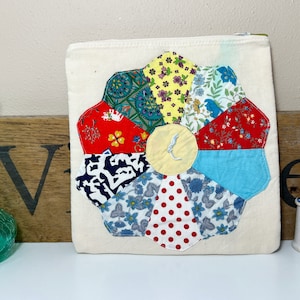 May include: A white canvas tote bag with a patchwork flower design. The flower is made of different fabrics in shades of red, blue, yellow, and green. The flower is centered on the bag and has a yellow center.