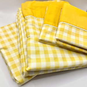 May include: A set of yellow and white gingham patterned bed sheets. The sheets have a solid yellow top with a white trim. The set includes a flat sheet, fitted sheet, and pillowcases. The bedding is folded and stacked.