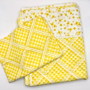 May include: A folded set of vintage bed sheets. One sheet features a yellow and white gingham pattern with a decorative lace trim. The other sheet has a white background with a pattern of small yellow flowers.