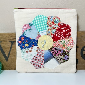 May include: A white canvas zippered pouch with a patchwork flower design. The flower is made of various fabrics in red, blue, green, and white with a yellow center.
