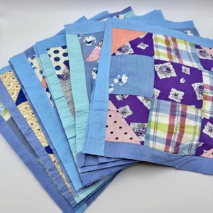May include: A stack of handmade patchwork squares in various colors and patterns. Each square features a light blue border and a center composed of fabric scraps in shades of blue, purple, pink, and white. The patchwork includes floral, polka dot, and plaid designs.