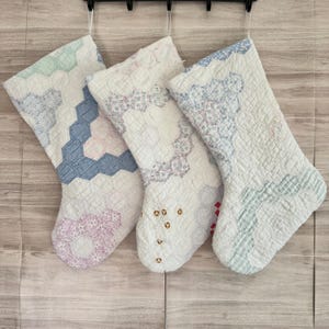 May include: Three quilted Christmas stockings with various pastel patterns, including hexagons and floral designs. The stockings are white with accents of blue, pink, and green. They hang from a black hook, showcasing a vintage, handmade aesthetic.