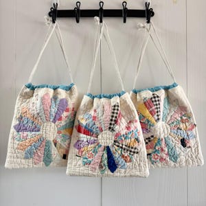 May include: Three quilted drawstring bags hanging from a black wall-mounted rack. Each bag features a cream-colored base with a large, multi-colored fabric flower appliqué made from various patterned textile strips and a central circular patch.