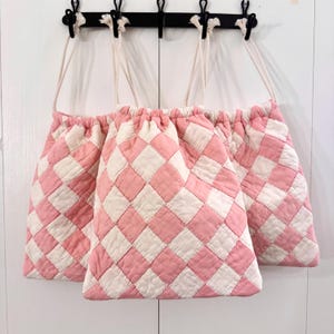 May include: Three pink and white checkered drawstring bags hanging on a black metal hook rack. The bags are made of quilted fabric and have white drawstrings. The bags are hanging against a white background.