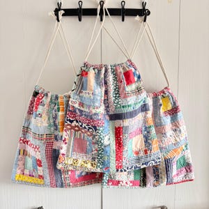 May include: Three patchwork drawstring bags with white rope straps hang from a black metal rack. The bags are made of various fabric scraps in a mix of colors and patterns, including red, blue, and floral prints. The bags are hanging against a white background.