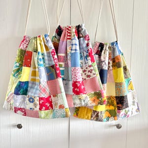 May include: Three drawstring bags made from patchwork fabric, each with a unique mix of patterns and colors. The bags hang from white cords against a white background. The patchwork includes floral, striped, and solid fabric pieces.
