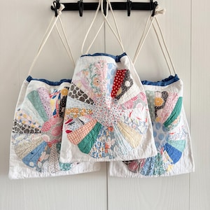 May include: Three white drawstring bags with a colorful patchwork design. Each bag features a fan-shaped pattern in various fabrics, including floral, polka dot, and geometric prints. The bags have a blue trim and white drawstring cords.
