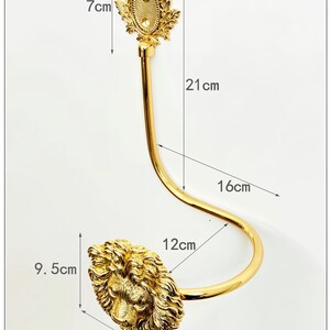 K&ouml;nnte beinhalten: Ein goldfarbener Wandhaken mit einem dekorativen L&ouml;wenkopf und einem verzierten Oberteil. Der L&ouml;wenkopf ist 9,5 cm breit und hoch. Der Haken ist 16 cm lang und 12 cm breit. Das Oberteil ist 7 cm breit und hoch.