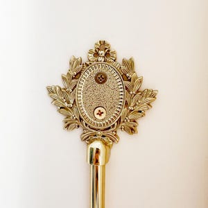 K&ouml;nnte beinhalten: Ein goldfarbener Gardinenstangen-Abschluss mit einem verzierten, blattartigen Design, das ein ovales Zentrum umgibt. Die Mitte hat eine strukturierte Oberfl&auml;che und ist mit zwei Schrauben befestigt. Die Stange selbst ist goldfarben poliert.