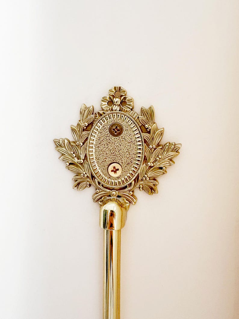 K&ouml;nnte beinhalten: Ein goldfarbener Gardinenstangen-Abschluss mit einem verzierten, blattartigen Design, das ein ovales Zentrum umgibt. Die Mitte hat eine strukturierte Oberfl&auml;che und ist mit zwei Schrauben befestigt. Die Stange selbst ist goldfarben poliert.