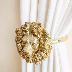K&ouml;nnte beinhalten: Ein goldfarbener Vorhanghalter in Form eines L&ouml;wenkopfes. Die M&auml;hne und die Gesichtsz&uuml;ge des L&ouml;wen sind aufwendig detailliert. Der Halter h&auml;lt wei&szlig;e Vorh&auml;nge. Der Hintergrund ist hellbeige.