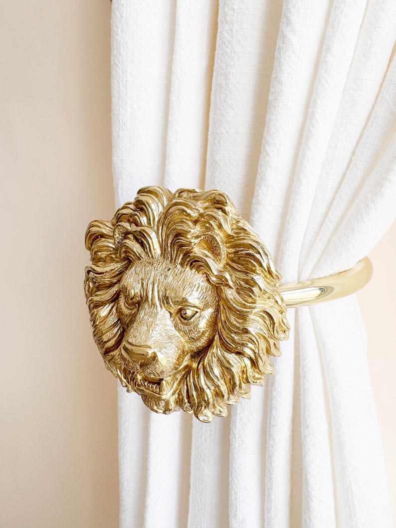 K&ouml;nnte beinhalten: Ein goldfarbener Vorhanghalter in Form eines L&ouml;wenkopfes. Die M&auml;hne und die Gesichtsz&uuml;ge des L&ouml;wen sind aufwendig detailliert. Der Halter h&auml;lt wei&szlig;e Vorh&auml;nge. Der Hintergrund ist hellbeige.
