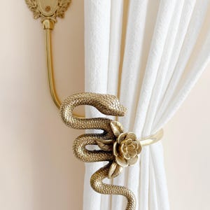 Può includere: Ferma tenda in metallo dorato con un design a serpente arrotolato e un accento floreale. Il fermatenda è montato su una parete con tende bianche sullo sfondo. È visibile anche un gancio a muro abbinato.