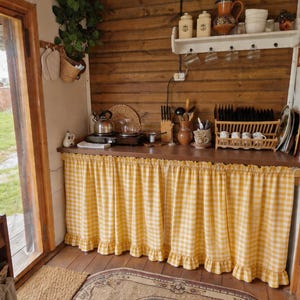 Può includere: Una scena accogliente della cucina con pareti in legno e un piano di lavoro coperto da una tenda a scacchi gialla e bianca. La cucina presenta una mensola con barattoli, tazze e una teiera. Un tappeto e una porta sono visibili.