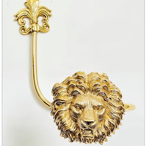 K&ouml;nnte beinhalten: Ein goldfarbener, dekorativer Gardinenhalter mit einem detaillierten L&ouml;wenkopf und einem Fleur-de-Lis-Design. Die M&auml;hne des L&ouml;wen ist aufwendig geformt, und der Halter hat einen gebogenen Arm. Der Hintergrund ist schlicht wei&szlig;.