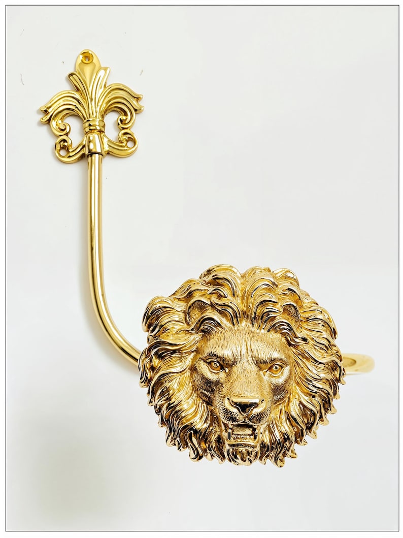 K&ouml;nnte beinhalten: Ein goldfarbener, dekorativer Gardinenhalter mit einem detaillierten L&ouml;wenkopf und einem Fleur-de-Lis-Design. Die M&auml;hne des L&ouml;wen ist aufwendig geformt, und der Halter hat einen gebogenen Arm. Der Hintergrund ist schlicht wei&szlig;.