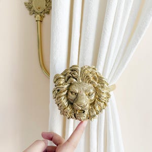 K&ouml;nnte beinhalten: Ein goldfarbener L&ouml;wenkopf-Vorhanghalter mit detaillierter M&auml;hne und offenem Maul, der wei&szlig;e Vorh&auml;nge h&auml;lt. Ein passender, an der Wand befestigter Gardinenstangenhalter ist ebenfalls sichtbar.