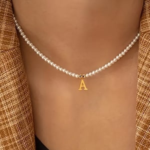 Peut inclure: Un collier de perles délicat avec un pendentif initial doré en forme de lettre "A". Le collier est porté sur une veste de couleur marron.
