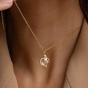 May include: A gold-colored necklace with a heart-shaped pendant. The pendant features a stylized letter "A" and a floral design. The necklace is worn around the neck and is displayed against a neutral background.