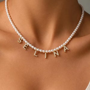 May include: A necklace featuring a strand of white pearls with gold-toned letter charms spelling the name SELINA. Each letter charm is encrusted with small clear stones, creating a sparkling effect against the smooth, round white pearls.